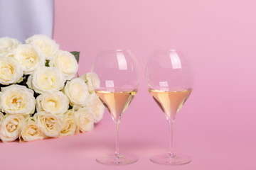 Beautiful romantic composition with white roses and two glasses of white wine on pink background. French white wine for romantic dinner. Holiday card with flowers and wine. Happy Valentine's Day