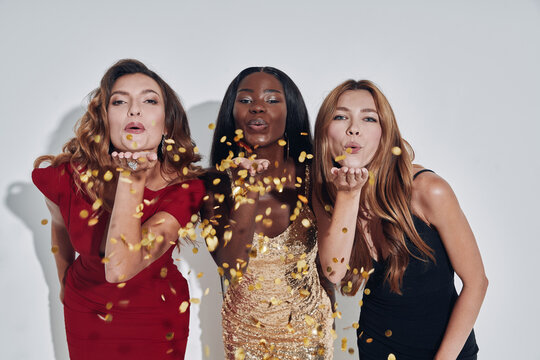 Three Beautiful Women In Evening Gowns Blowing Confetti And Smiling While Standing Against Gray Background