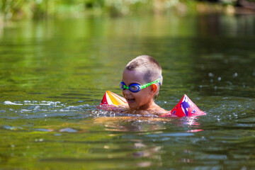 Cute boy is swimming in the small river with his armbands and goggles