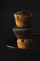 Delicious vanilla and chocolate muffins with chocolate chips next to a cup of coffee and a black cover book on a black background on a dark wooden table
