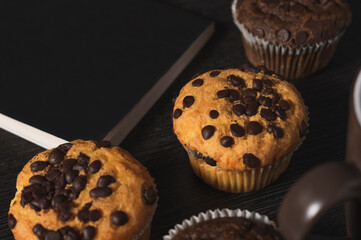Delicious vanilla and chocolate muffins with chocolate chips next to a cup of coffee and a black cover book on a black background on a dark wooden table