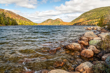 Two granite mountain peaks, called The Bubbles, in Acadia National Park, are bathed in sunlight and autumn foliage color across Jordan Pond on a partly cloudy fall day in Down East Maine.