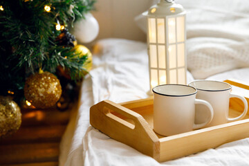 Fototapeta na wymiar wooden tray and two white mugs on the bed on a white sheet, near a decorated Christmas tree. Cozy morning. Coffee in bed