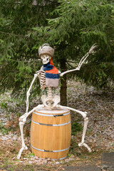 A skeleton in a winter hat and scarf sits on a wooden barrel under a fir tree.
