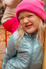 Cute funny blond preschool girl making faces outdoors on a autumn day. Girl wearing pink beanie and blue wind jacket. Vertical shot