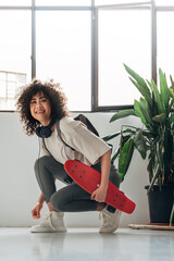Smiling young multiracial woman tying sneaker shoelace while holding skateboard and wearing...