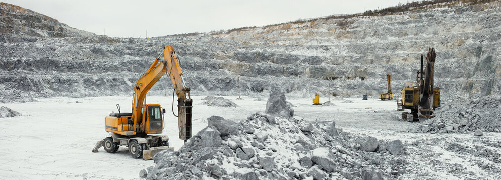 Panorama of a limestone quarry with heavy mining machinery - hydraulic hammer, excavator and drilling rig, on a cloudy winter day.
