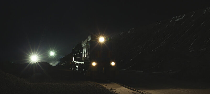 Silhouette of a diesel locomotive is moving on railroad tracks in the dark with artificial lighting of searchlights at a mining plant, panorama.