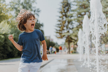 little boy is running in the street and playing with the water jets of a fountain spouting from the...