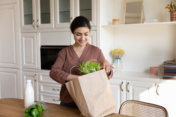Smiling Indian woman put paper shopping bag full with fresh quality groceries products on kitchen table unpack dairy vegetables purchased on-line, supermarket food delivery, comfort services concept