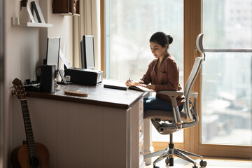 Serious young Indian female sit on ergonomic comfy chair at desk holds pen jotting information,...