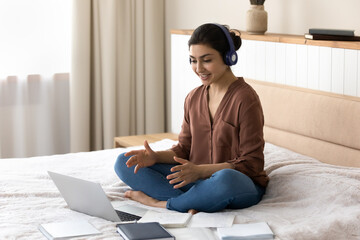 Indian student woman sit on bed wear headphones talk on video conference, e-learning preparing for...