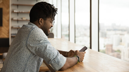 Smiling young Brazilian ethnicity man looking at cellphone screen, reading message with good news, playing online game, shopping online or web surfing in modern office, tech addiction.