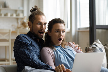 Shocked young bonding man woman family couple looking at laptop screen, reading email with unexpected news, surprised by unbelievable opportunity, online discount sale, sitting on sofa at home.