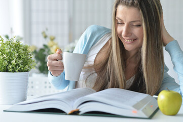 Beautiful young woman with cup of tea and reading book at home