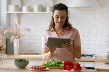 Happy young beautiful woman using digital computer tablet, enjoying communicating in social network, web surfing recipes online or checking messages, cooking healthy food alone in modern kitchen.