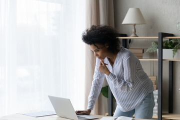 Pensive African American woman standing near work desk at home, using laptop, touching chin and...