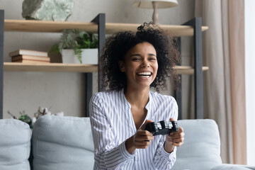 Laughing overjoyed African American woman playing video game, sitting on couch at home, happy...