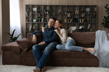 Obraz premium Happy dreamy Caucasian loving couple hugging, relaxing sitting on couch at home, smiling wife and husband looking to aside, dreaming of good future, new opportunities together, enjoying weekend