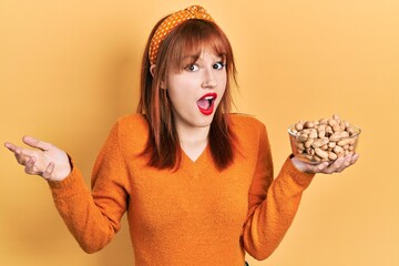 Redhead young woman holding peanuts celebrating achievement with happy smile and winner expression with raised hand