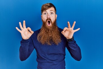 Redhead man with long beard wearing casual blue sweater over blue background looking surprised and shocked doing ok approval symbol with fingers. crazy expression