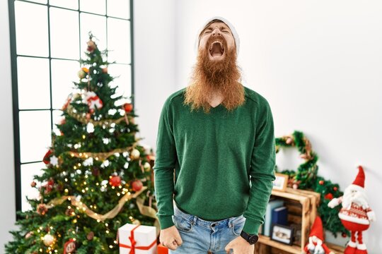 Redhead man with long beard wearing christmas hat by christmas tree angry and mad screaming frustrated and furious, shouting with anger. rage and aggressive concept.