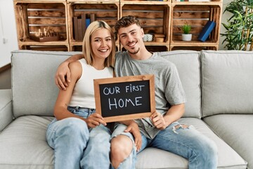 Young caucasian couple smiling happy holding blackboard with our first home message sitting on the sofa. - obrazy, fototapety, plakaty