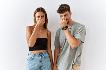Young beautiful couple standing together over isolated background smelling something stinky and disgusting, intolerable smell, holding breath with fingers on nose. bad smell - obrazy, fototapety, plakaty