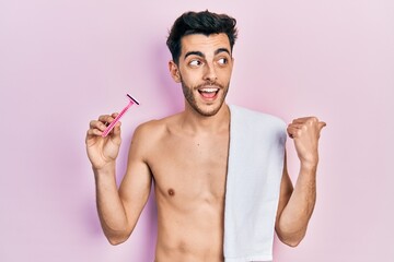 Young hispanic man standing shirtless holding razor pointing thumb up to the side smiling happy with open mouth