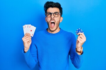 Young hispanic man playing poker holding casino chips and cards angry and mad screaming frustrated...