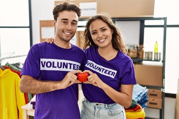Young hispanic couple wearing volunteer t shirt holding heart smiling with a happy and cool smile on face. showing teeth.