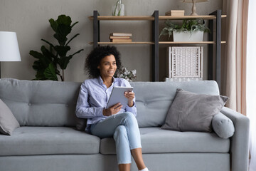 Fototapeta premium Smiling thoughtful African American woman holding tablet, looking in distance, happy dreamy young female distracted from online chatting, visualizing good future, sitting on cozy couch at home