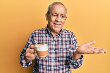 Handsome senior man with grey hair drinking a cup coffee smiling cheerful presenting and pointing with palm of hand looking at the camera.