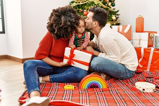 Couple and daughter holding gift and kissing sitting by christmas tree at home