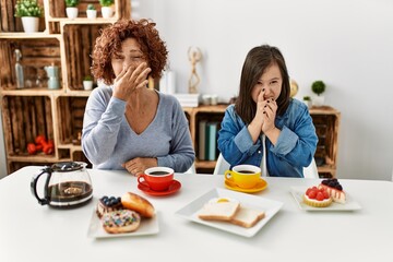 Family of mother and down syndrome daughter sitting at home eating breakfast smelling something stinky and disgusting, intolerable smell, holding breath with fingers on nose. bad smell - obrazy, fototapety, plakaty