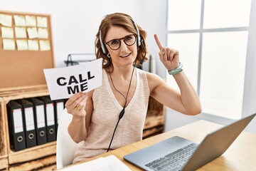 Middle age brunette woman wearing operator headset holding call me banner smiling amazed and...