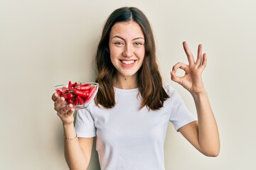 Young brunette woman holding red peppers doing ok sign with fingers, smiling friendly gesturing excellent symbol