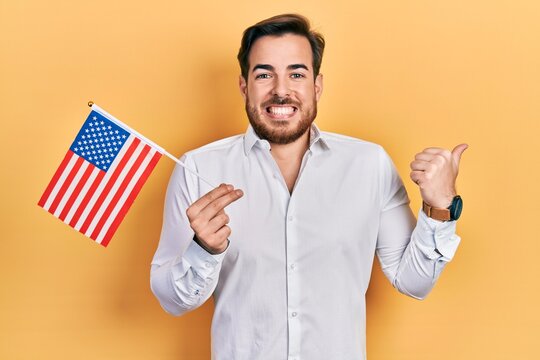 Handsome caucasian man with beard holding united states flag pointing thumb up to the side smiling happy with open mouth