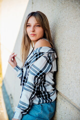 Fototapeta premium A young girl in a plaid shirt is standing near a gray wall. A thoughtful and dreamy look to the side.