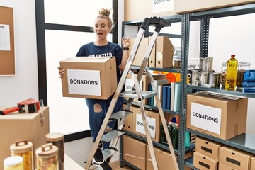 Young caucasian woman volunteer holding donations box with a big smile on face, pointing with hand finger to the side looking at the camera.