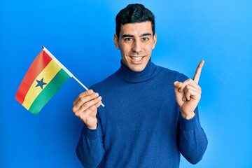 Handsome hispanic man holding ghana flag smiling happy pointing with hand and finger to the side