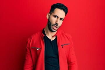 Young hispanic man wearing red leather jacket in shock face, looking skeptical and sarcastic, surprised with open mouth