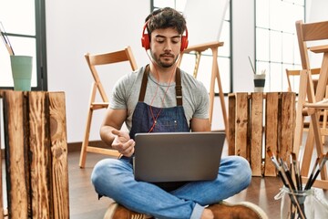 Young hispanic artist man using headphones and laptop at art studio.