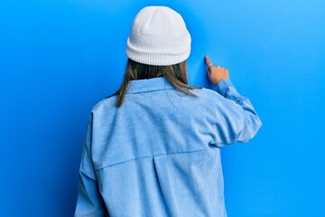 Young latin woman wearing cute wool cap posing backwards pointing ahead with finger hand