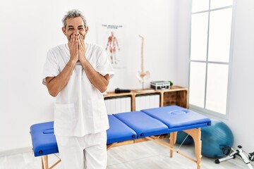 Middle age hispanic therapist man working at pain recovery clinic laughing and embarrassed giggle...