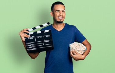 Young african american man eating popcorn holding film clapboard winking looking at the camera with...