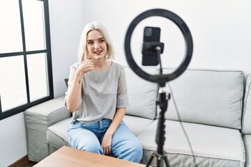 Fototapeta na wymiar Young caucasian woman recording vlog tutorial with smartphone at home doing happy thumbs up gesture with hand. approving expression looking at the camera showing success.