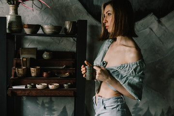 young and beautiful girl in blue blouse and light jeans is standing in a pottery workshop with a clay mug of tea. Concept hobby, applied art
