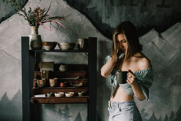 young and beautiful girl in blue blouse and light jeans is standing in a pottery workshop with a clay mug of tea. Concept hobby, applied art