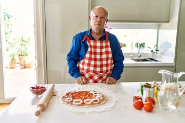 Senior man with grey hair cooking pizza at home kitchen with serious expression on face. simple and natural looking at the camera.